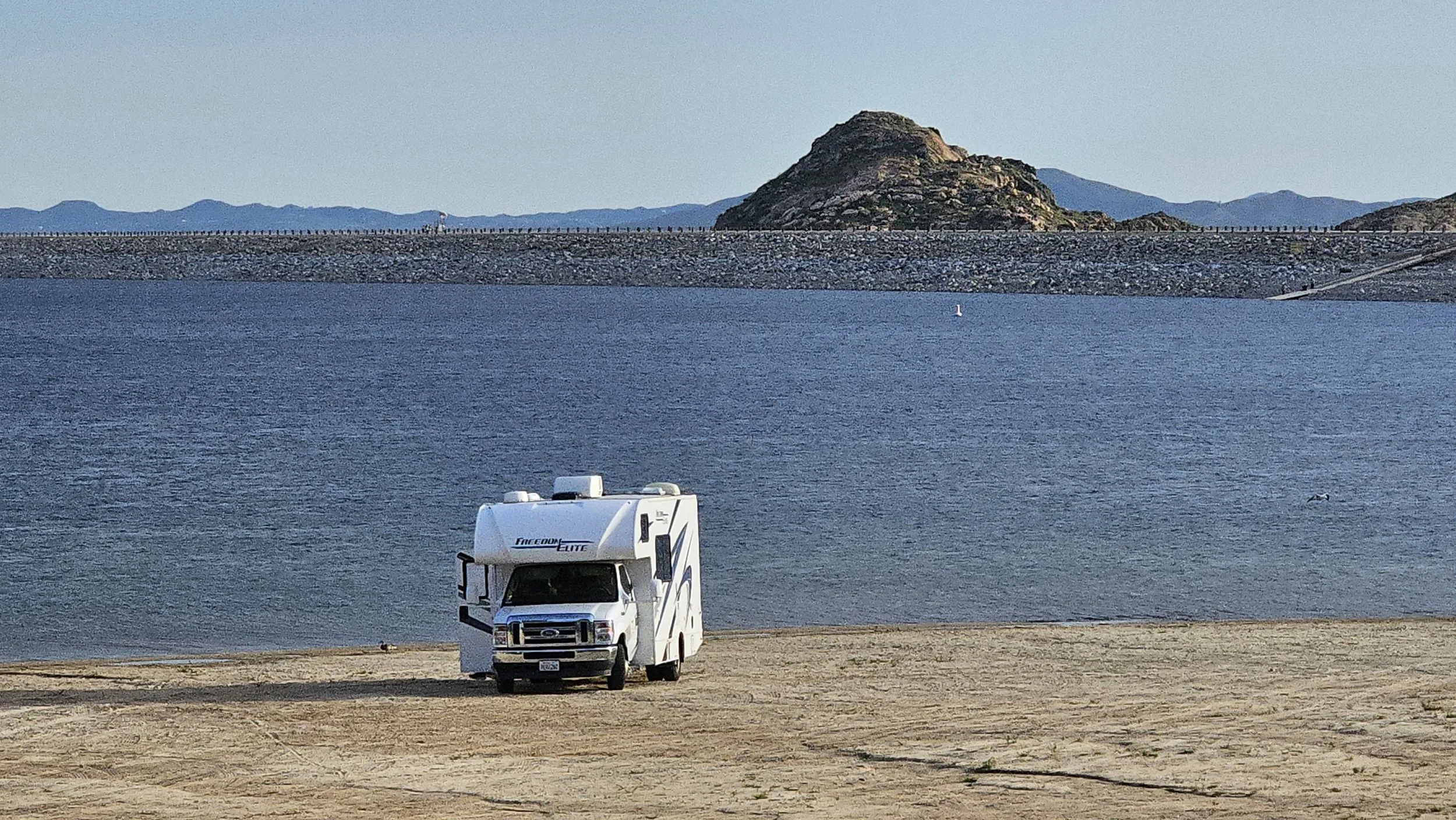 A recreational vehicle (RV) parked on a sandy beach by a large body of water, with a rocky hill and distant mountains in the background.