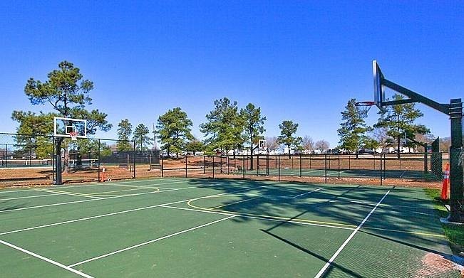 An empty outdoor basketball court with two hoops, surrounded by a fence, set against a backdrop of trees and a clear blue sky.
