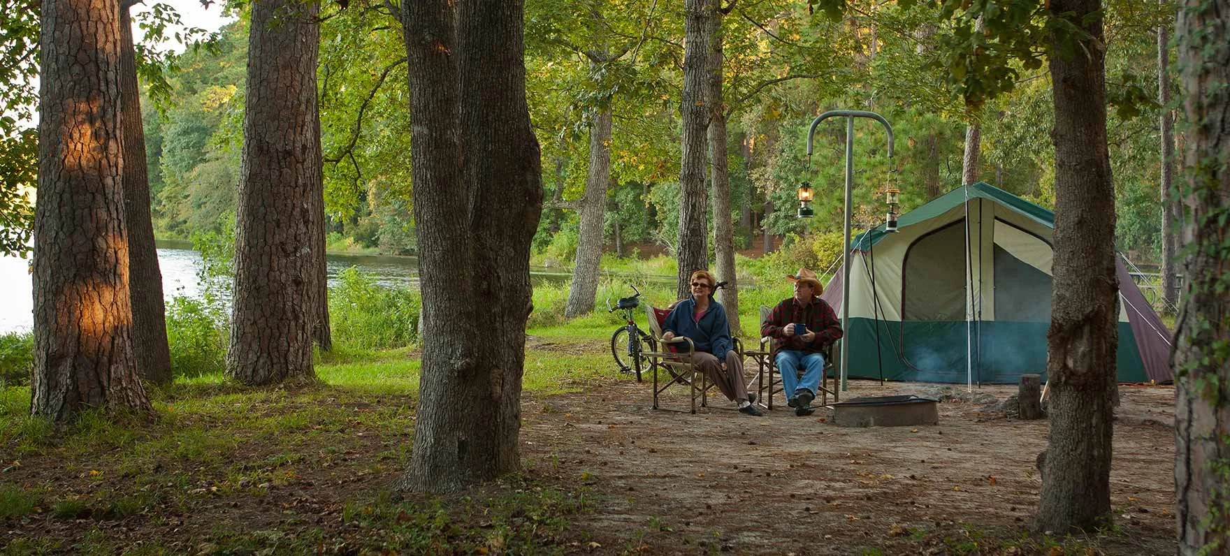 Two people sitting on camping chairs near a tent in a wooded campsite by a river, with a bicycle nearby and lanterns hanging, during daytime.