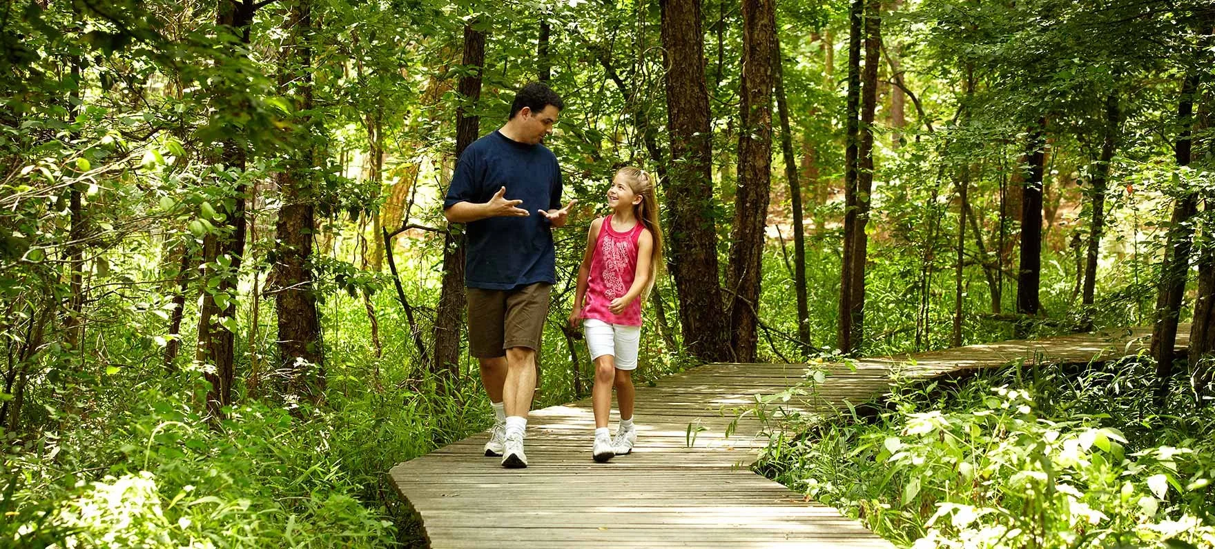 A man and a girl walking and talking on a wooden trail through a dense, green forest.