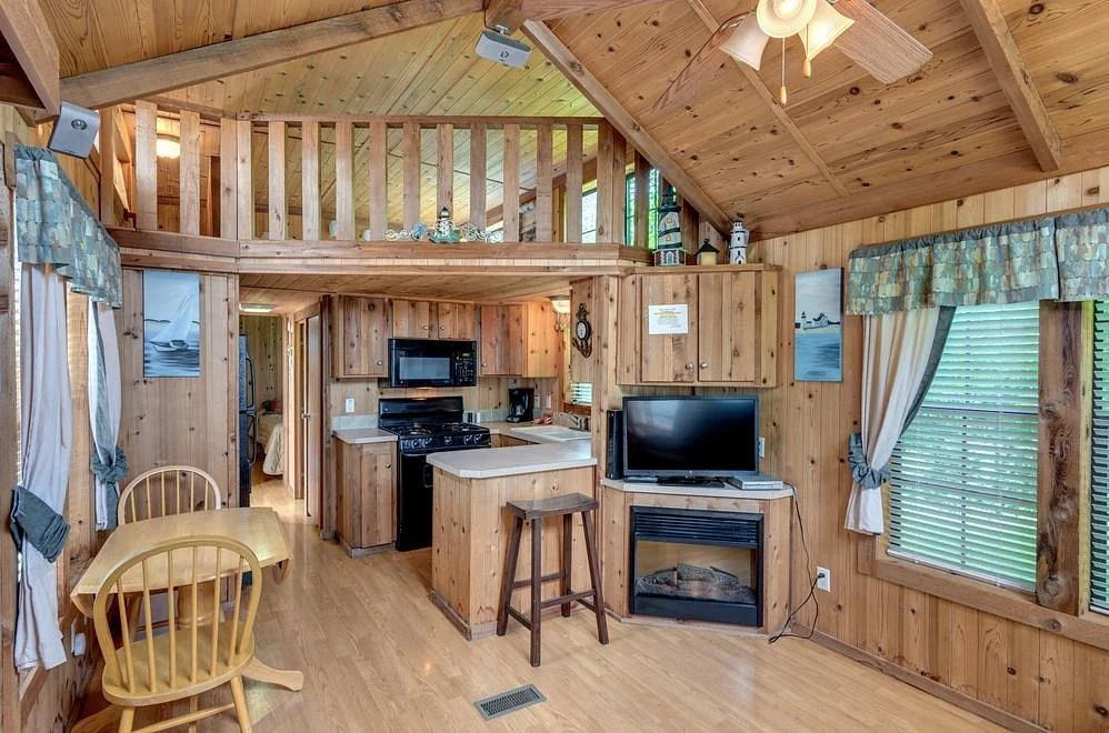 Interior of a cabin with wood-paneled walls and ceiling, featuring a small kitchen with black appliances, a TV on a stand, and a dining table with wooden chairs. The room has windows with curtains, and a loft area above with a railing.