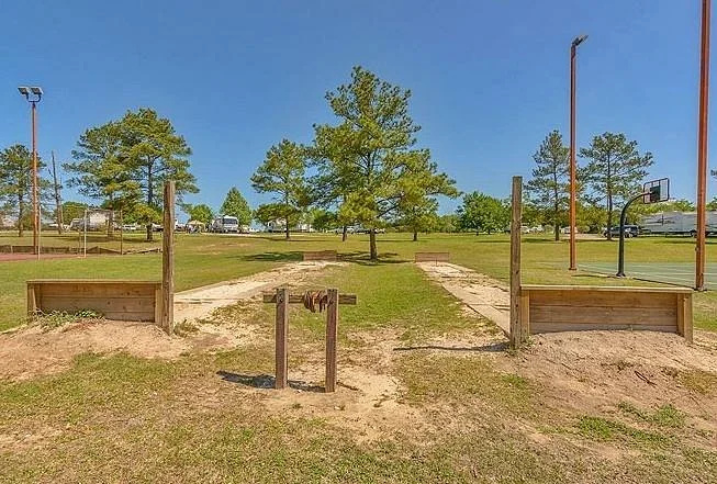 A playground area with two wooden sandbox structures, a jumping pole, and a baseball court with a hoop, surrounded by grassy fields and trees under a clear blue sky.