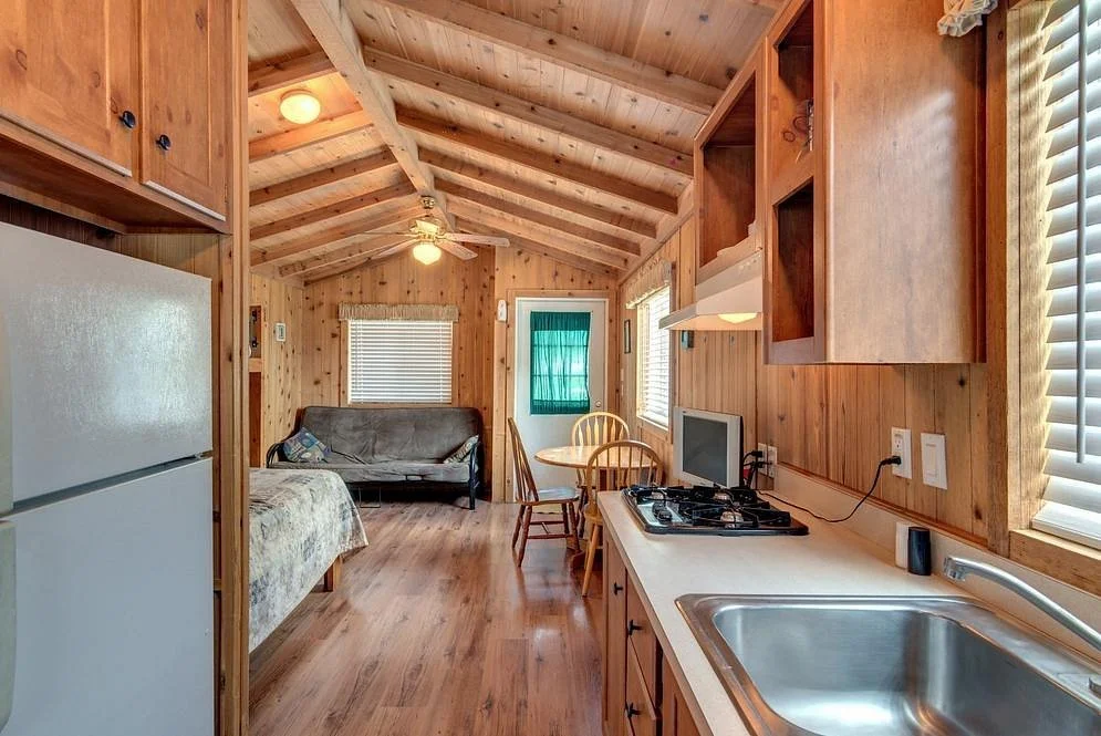Interior view of a small wooden cabin with kitchen, dining, and living area, featuring a refrigerator, stove, sink, chairs, and a sofa.