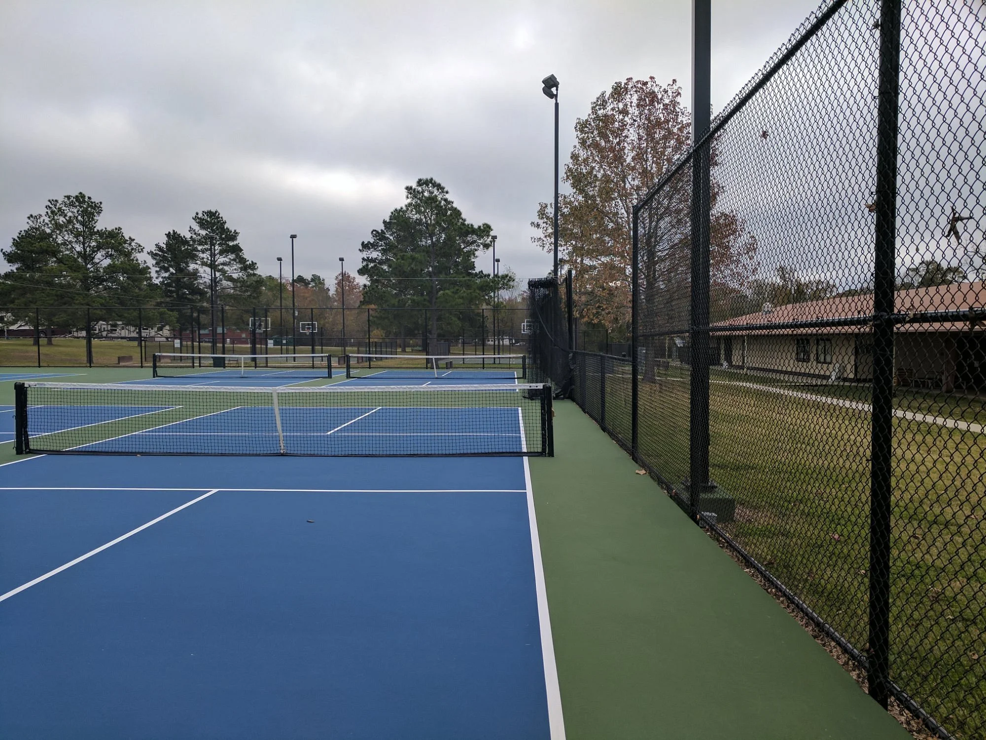 An outdoor tennis court with blue surface and white lines, surrounded by black chain-link fencing, under a cloudy sky.
