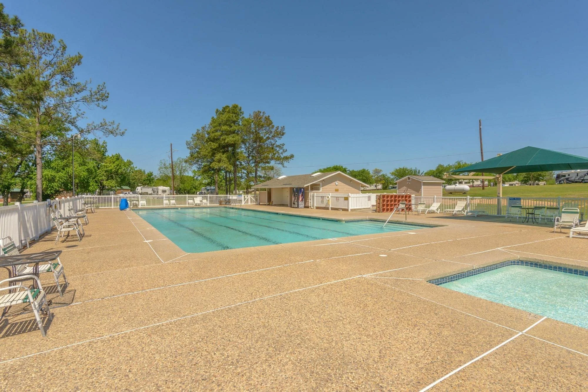 Empty outdoor swimming pool and hot tub at a recreational facility with clear blue sky, surrounded by white fence, trees, and poolside chairs with umbrellas.