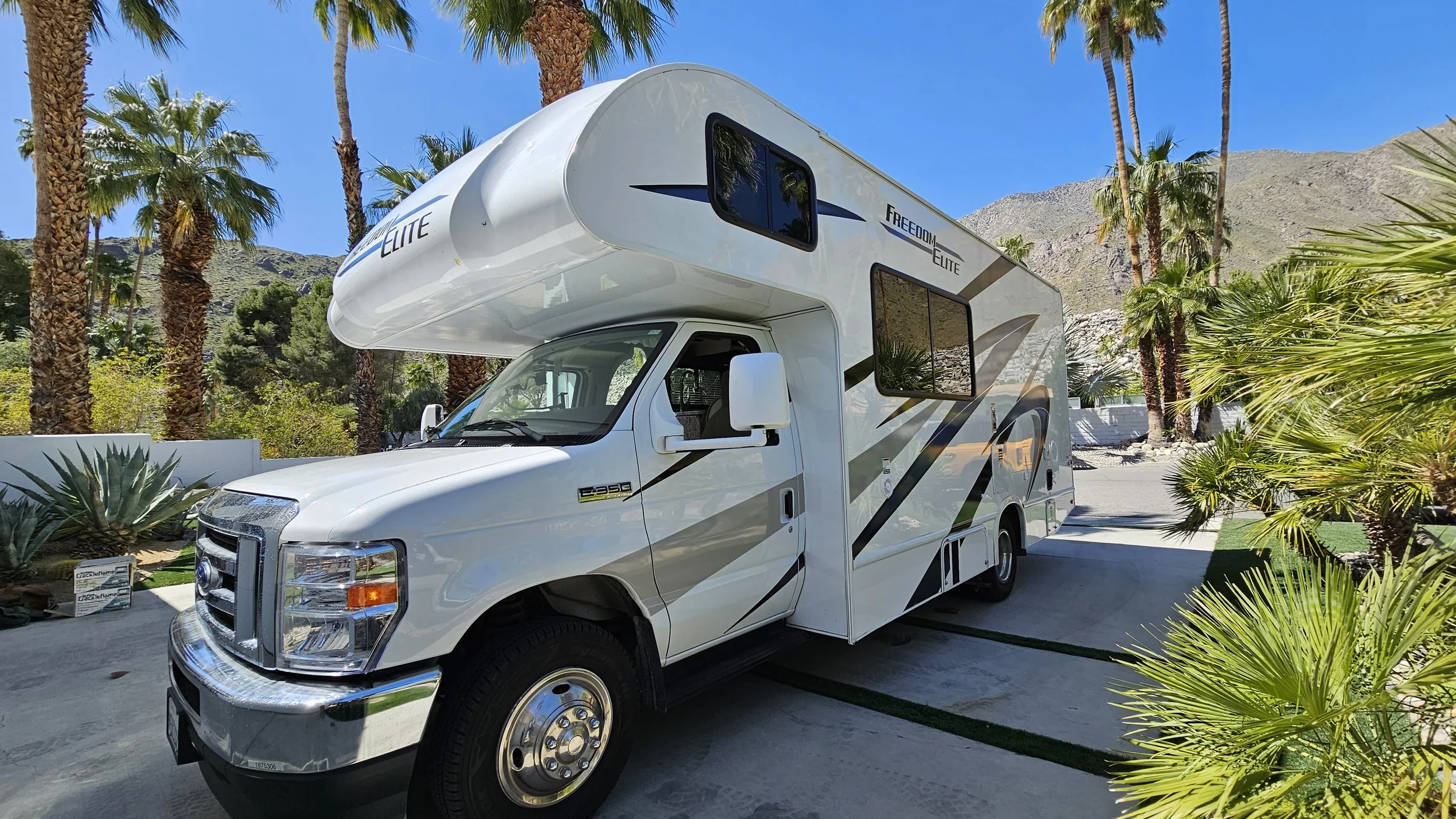 A white RV with the words 'FREEDOM ELITE' parked on a driveway surrounded by palm trees and desert landscaping under a clear blue sky.
