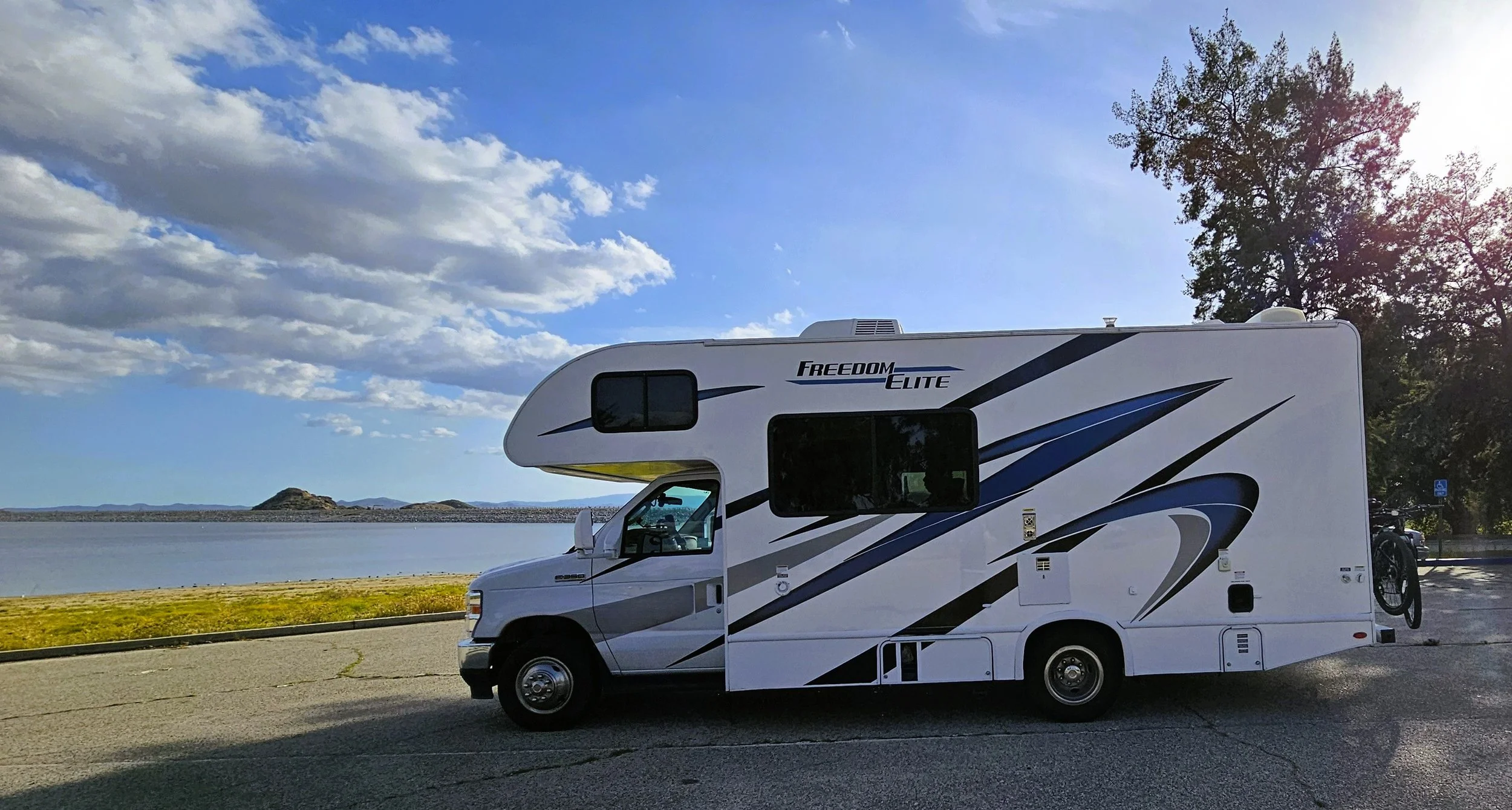 A white recreational vehicle (RV) with blue and black graphics, labeled 'Freedom Elite,' parked on a paved lot near a body of water. The sky is partly cloudy, and trees are visible in the background.