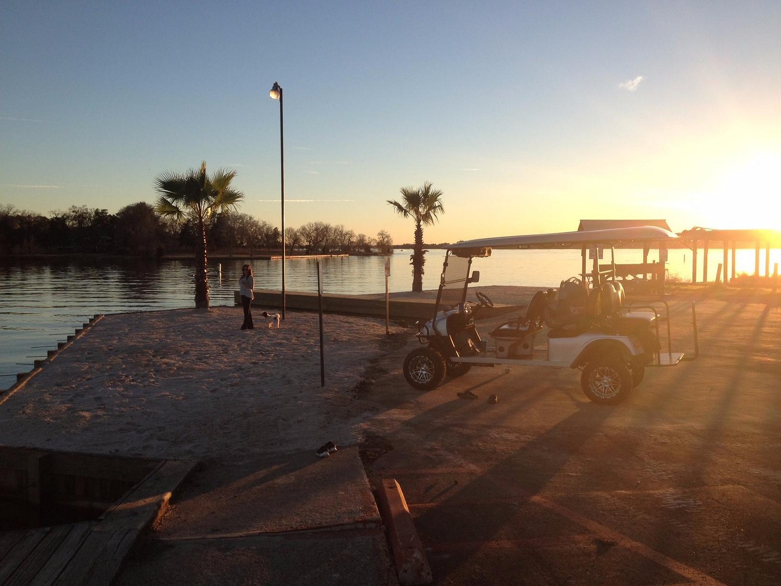 A waterfront scene during sunset with a person and a dog on the sandy beach, palm trees, a golf cart, and a pier in the background.