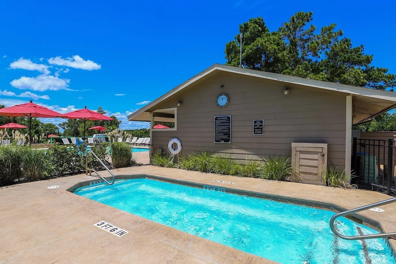 An outdoor swimming pool area with lounge chairs, red umbrellas, and a small pool for children, surrounded by greenery and under a bright blue sky.