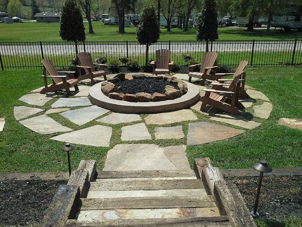 A circular outdoor firepit surrounded by six Adirondack chairs on a stone patio, with a small wooden staircase leading to the seating area, enclosed by a black metal fence with trees and a grassy yard in the background.