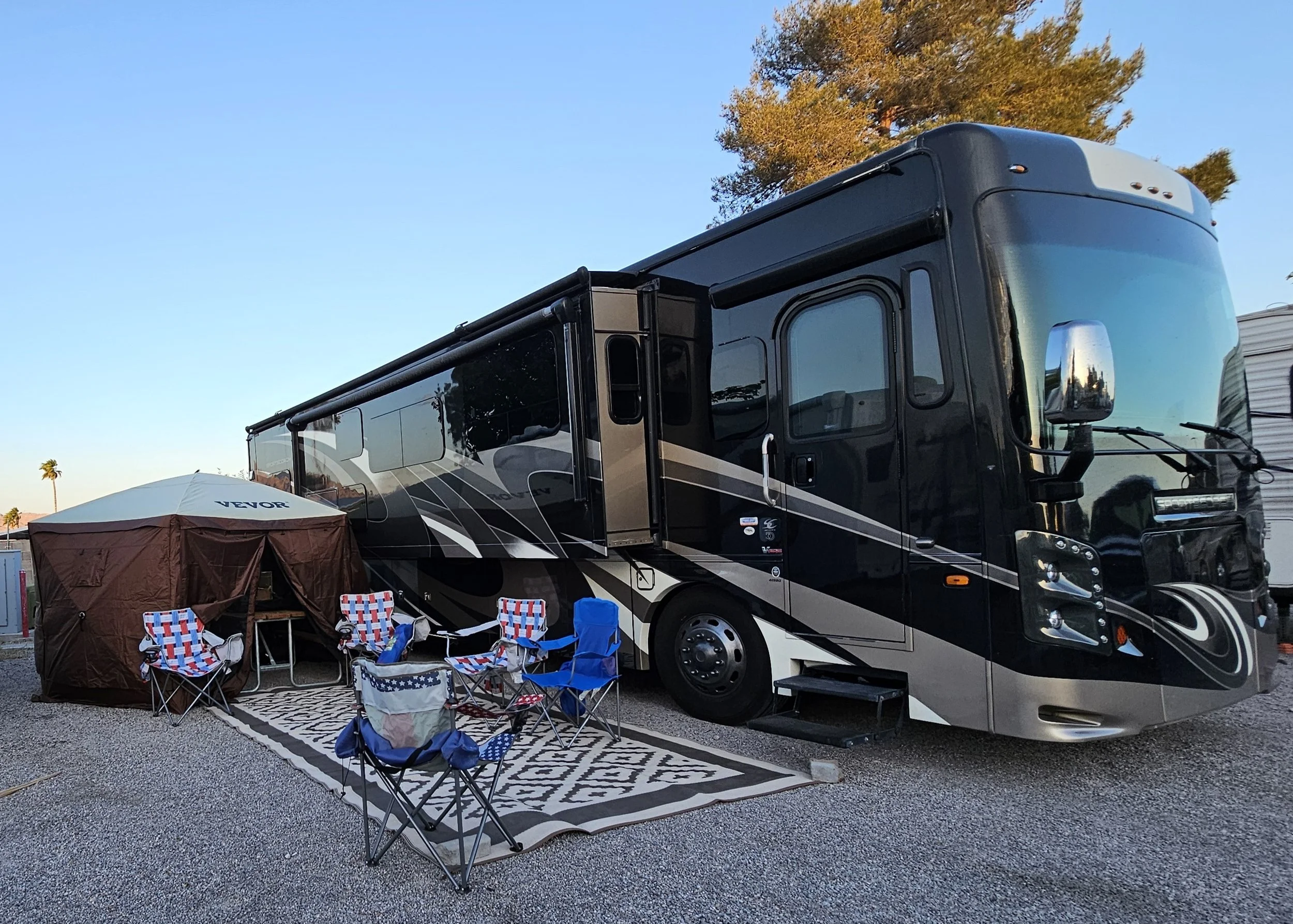 A black and gray RV parked on a gravel lot with outdoor chairs and a small tent, set up for camping, during sunset or early evening.