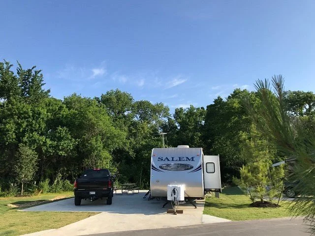 A parked Salem travel trailer hitched to a black pickup truck on a concrete pad, surrounded by green trees and bushes, with a clear blue sky overhead.