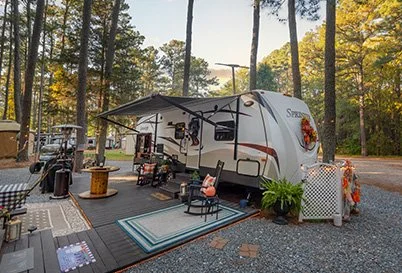 A recreational RV trailer parked in a wooded area with outdoor chairs, a table, a potted plant, and decorative fall pumpkins.