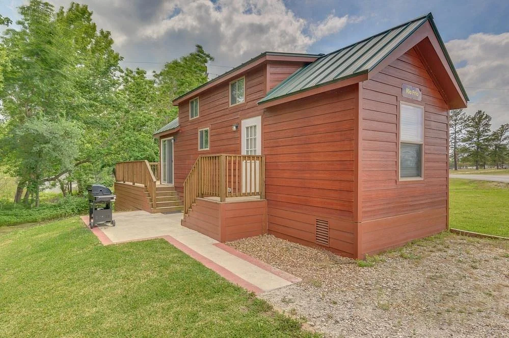 Red wooden house with a metal roof, front steps, small deck, and a backyard grill, surrounded by green grass and trees.