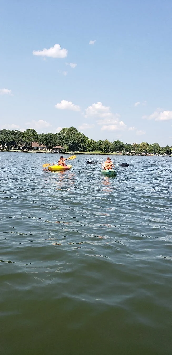 Two people kayaking on a lake under a partly cloudy sky, with trees and homes in the background.