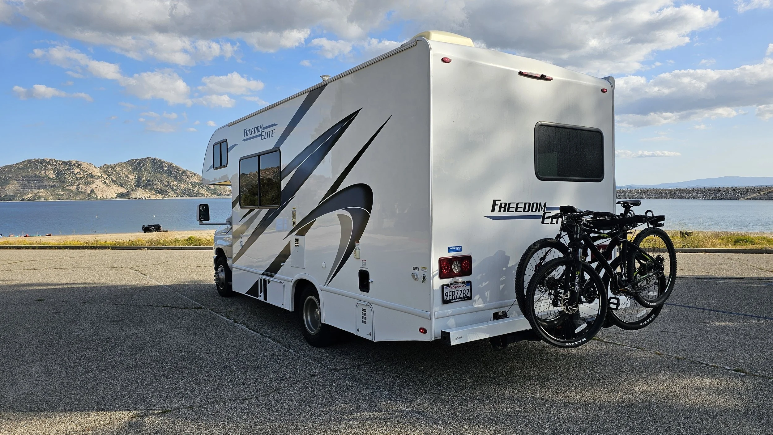A white motorhome with two bikes attached at the back, parked near a body of water with mountains in the background and a blue sky with clouds.