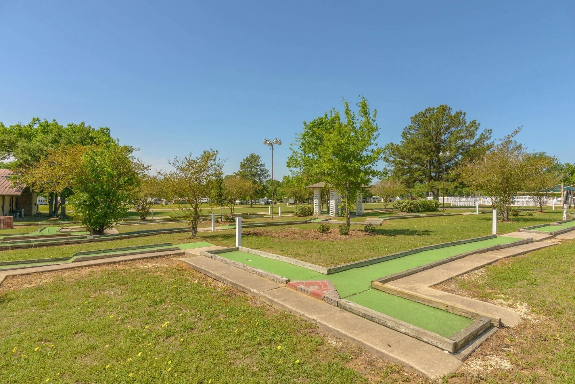 A miniature golf course with several green-painted holes, pathways, and goal areas, surrounded by a grassy area with trees and a small gazebo under a clear blue sky.