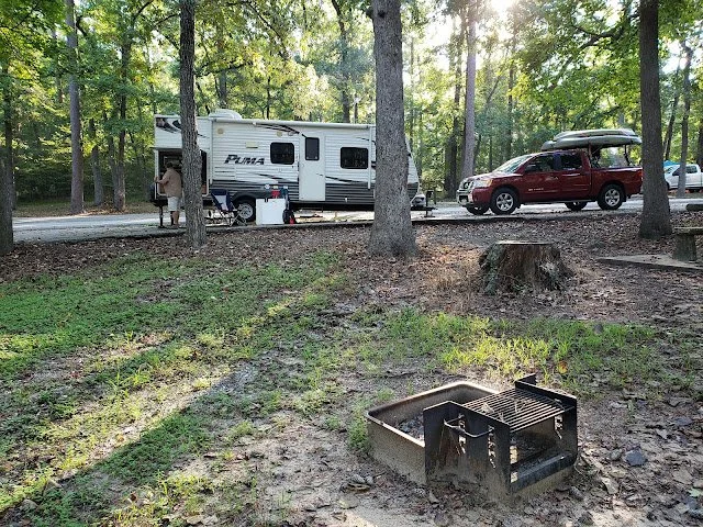 A campground scene with a white camper trailer, a red SUV with a kayak on top, a person standing near the camper, and a small non-functioning fire pit in the foreground surrounded by trees.