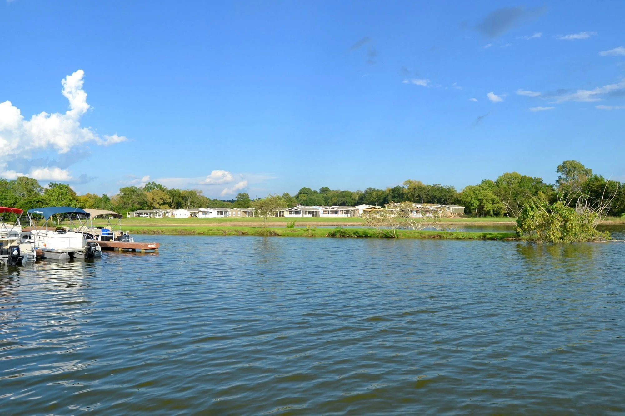 A view of a calm water body with boats docked on a marina on the left, an island with green bushes in the middle, and a line of houses and trees in the background under a partly cloudy blue sky.