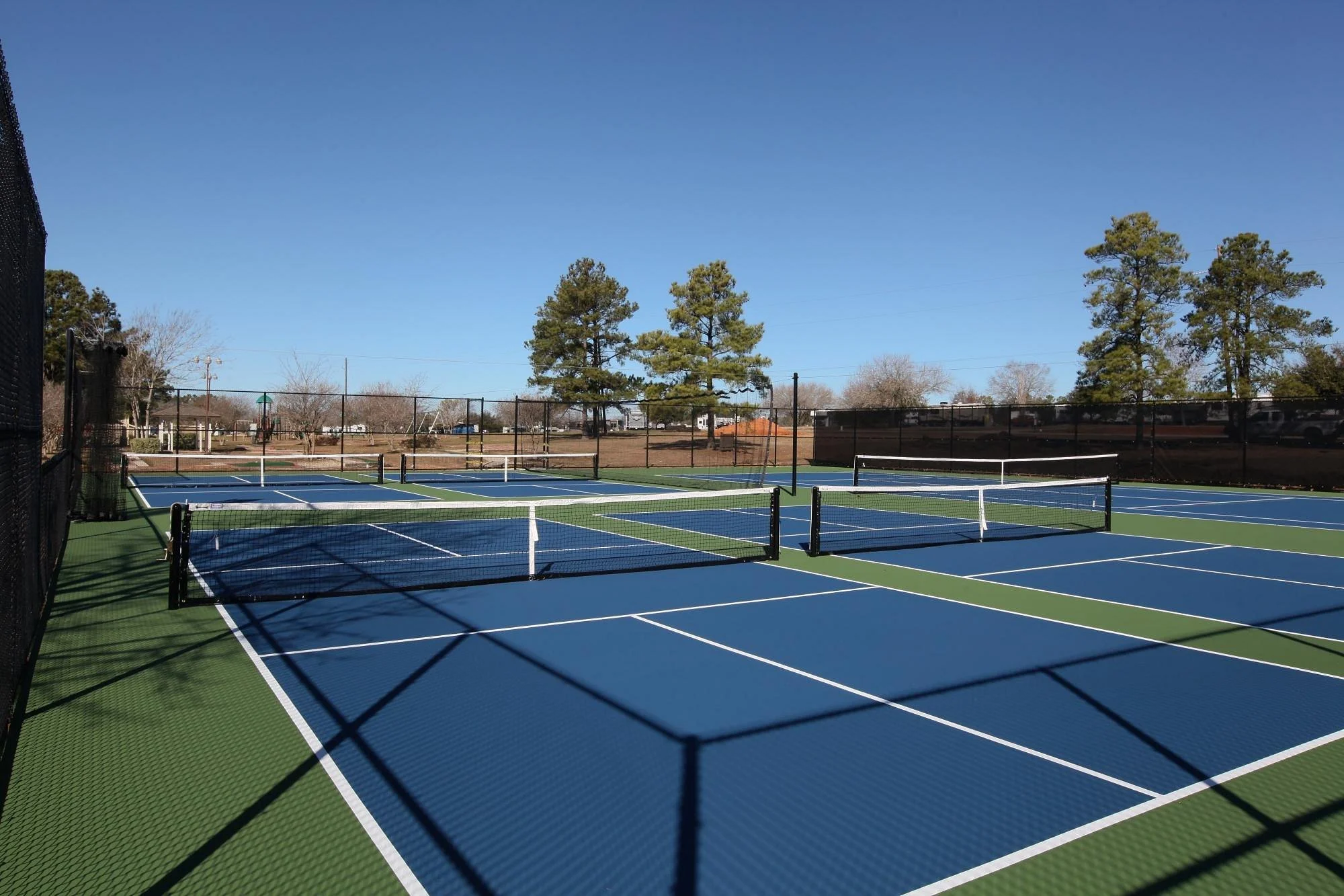 Multiple blue and green tennis courts with white lines, surrounded by black fences, under a clear blue sky with trees in the background.