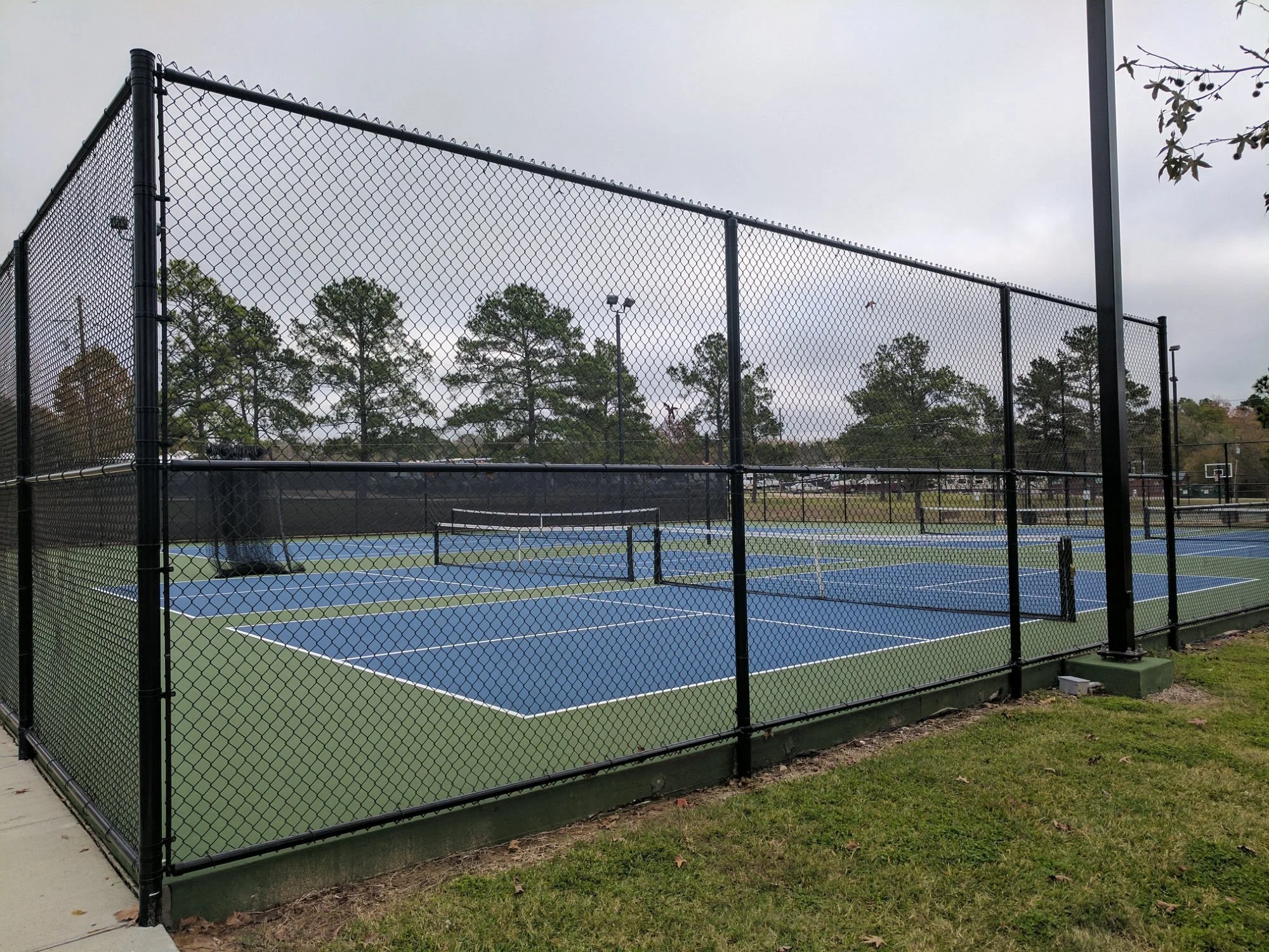 An outdoor tennis court with blue playing surface and a black chain-link fence surrounding it, with trees and a cloudy sky in the background.
