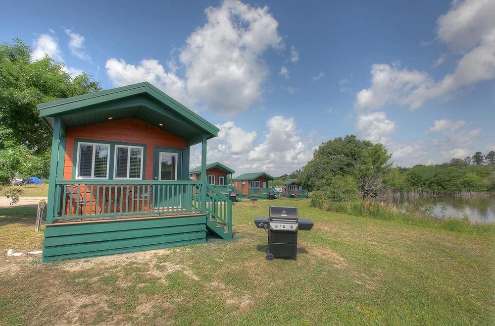 Small cabin with green trim, front porch, and stairs, next to a lake with grassy bank, trees, and a grill on the lawn under a partly cloudy sky.