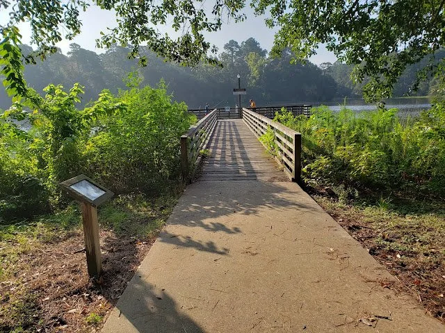 A wooden bridge extending over a body of water, surrounded by green trees and bushes, with a signboard on the left side and a person visible at the bridge's end.
