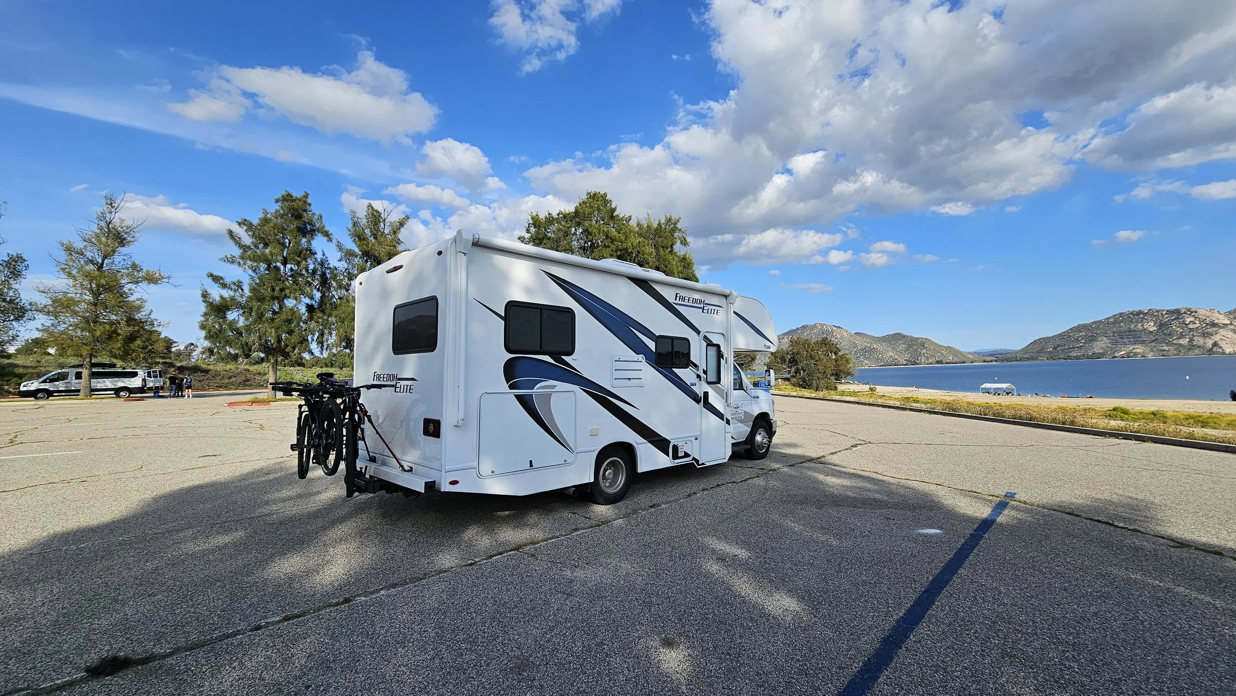 A white camper van with black and blue graphics parked in an open lot near a lake, with a bicycle attached at the back. The background features trees, water, hills, and a partly cloudy sky.