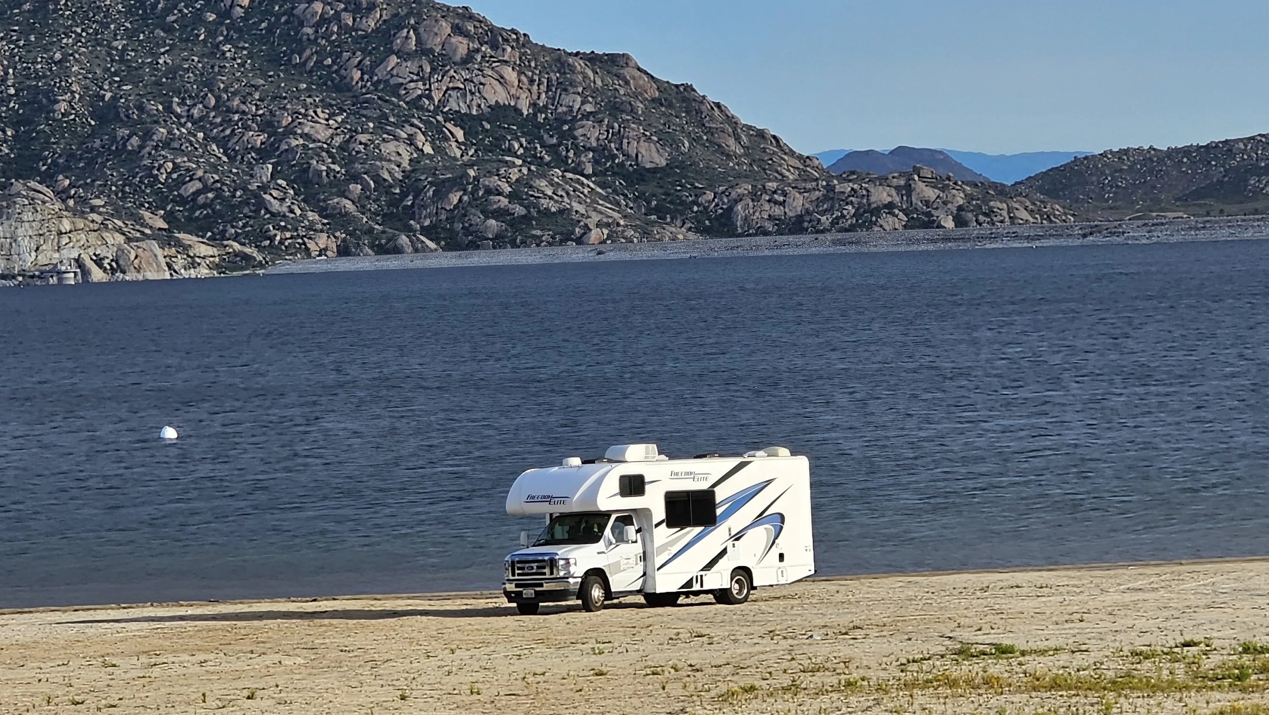 A white RV parked on a sandy area beside a large body of water, with mountains in the background under a clear blue sky.