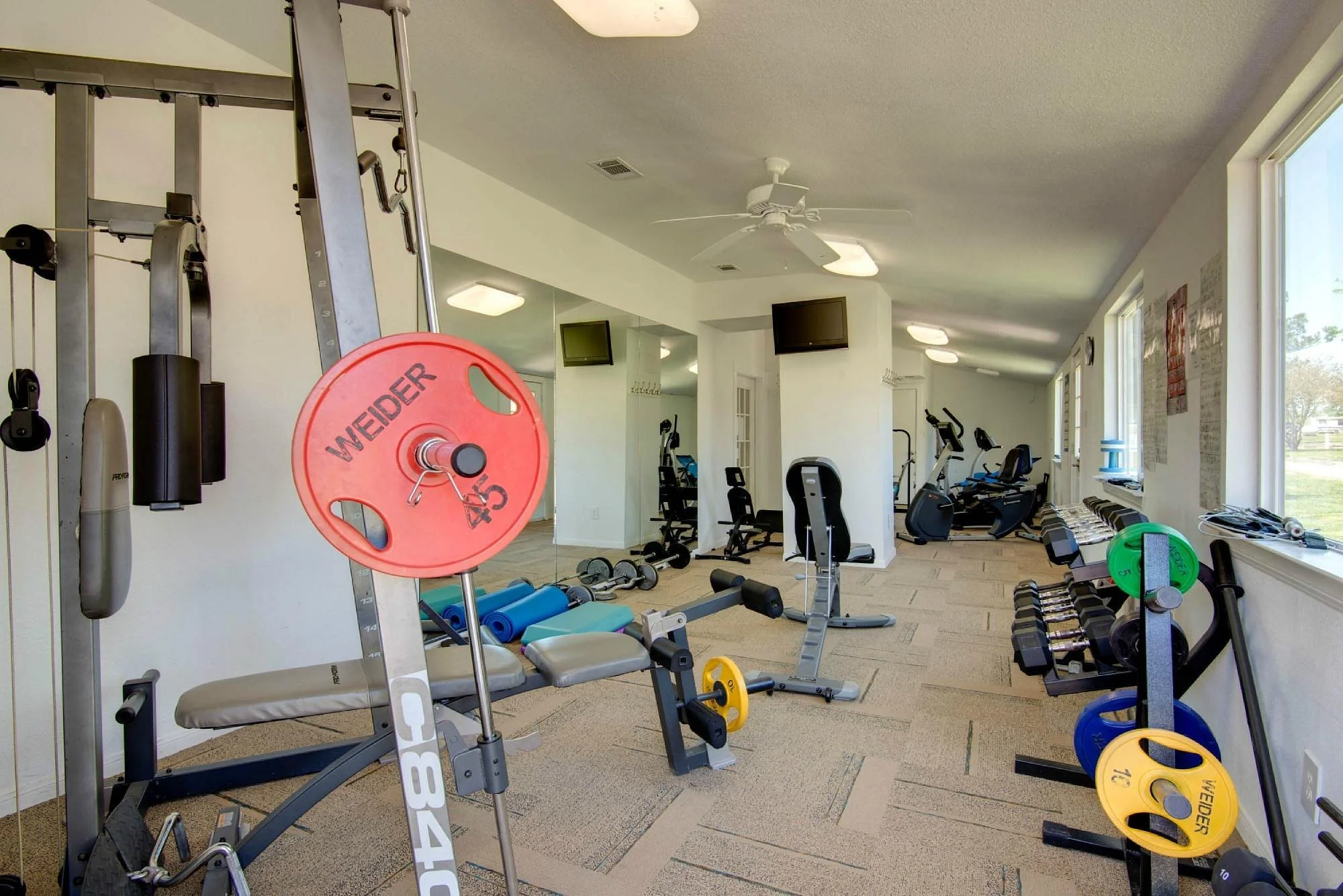 Well-lit fitness room with various exercise equipment including weights, benches, stationary bikes, and cardio machines, with windows on the right side and a ceiling fan overhead.