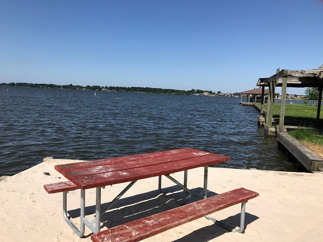 A red picnic table on a sandy beach near a body of water, with a wooden dock and a shoreline with houses in the background under a clear blue sky.