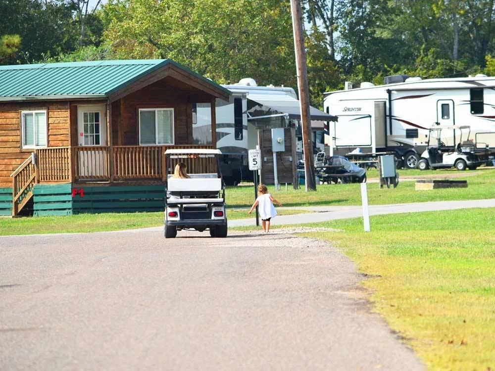 A young girl walking on a paved road towards a golf cart parked near a wooden cabin with a green metal roof, surrounded by RVs and trees in the background.