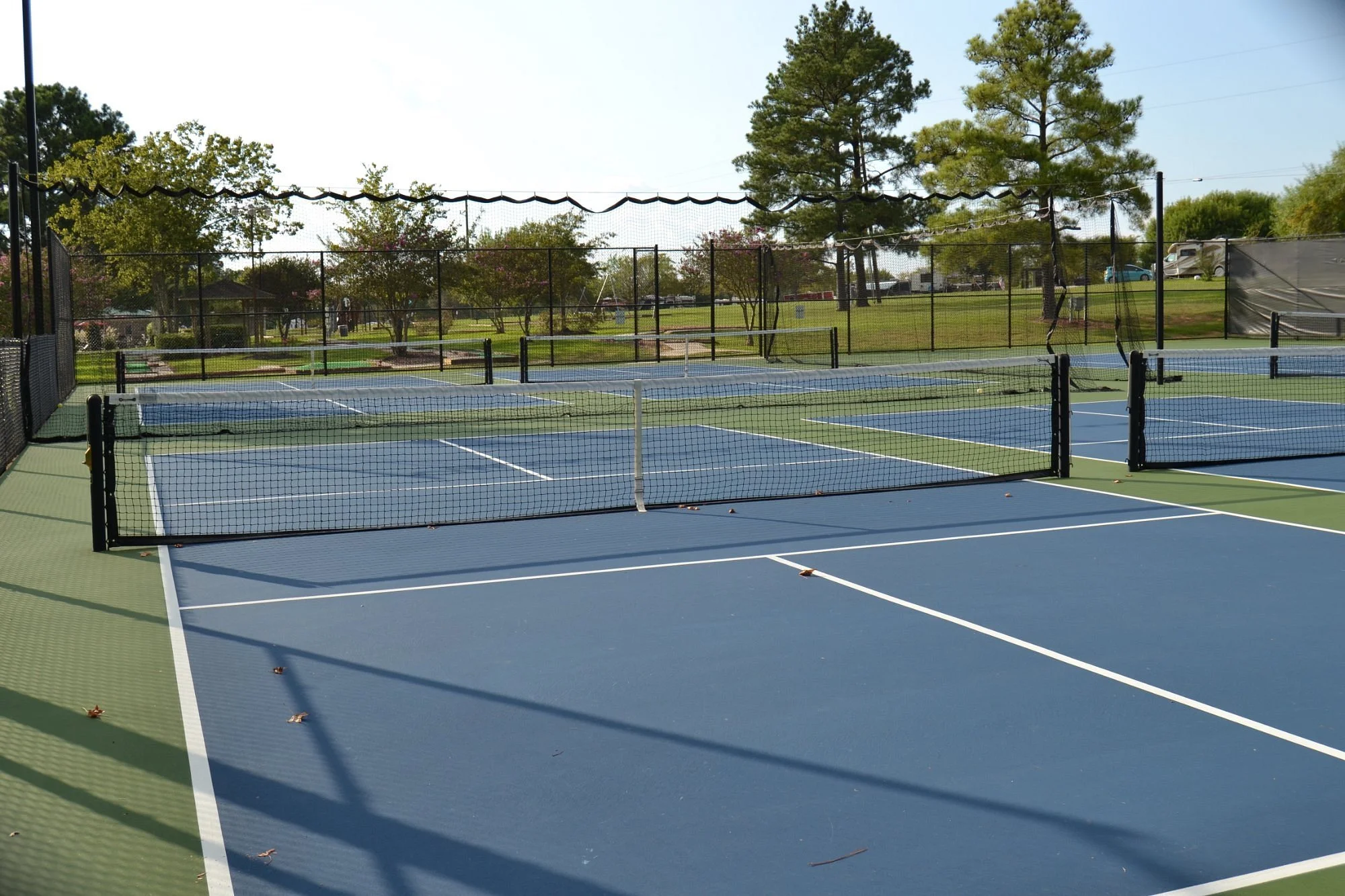 Empty outdoor tennis courts with blue and green surfaces, surrounded by black fencing and trees in the background.
