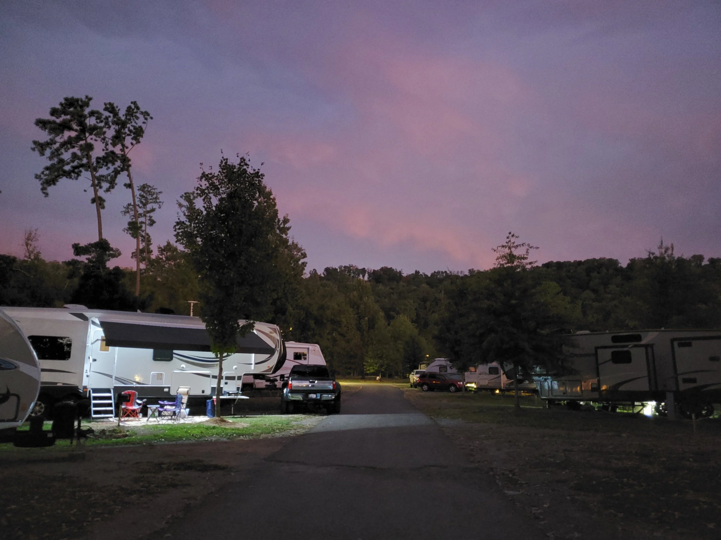 Camping area at dusk with several RVs parked, trees, and a colorful evening sky.