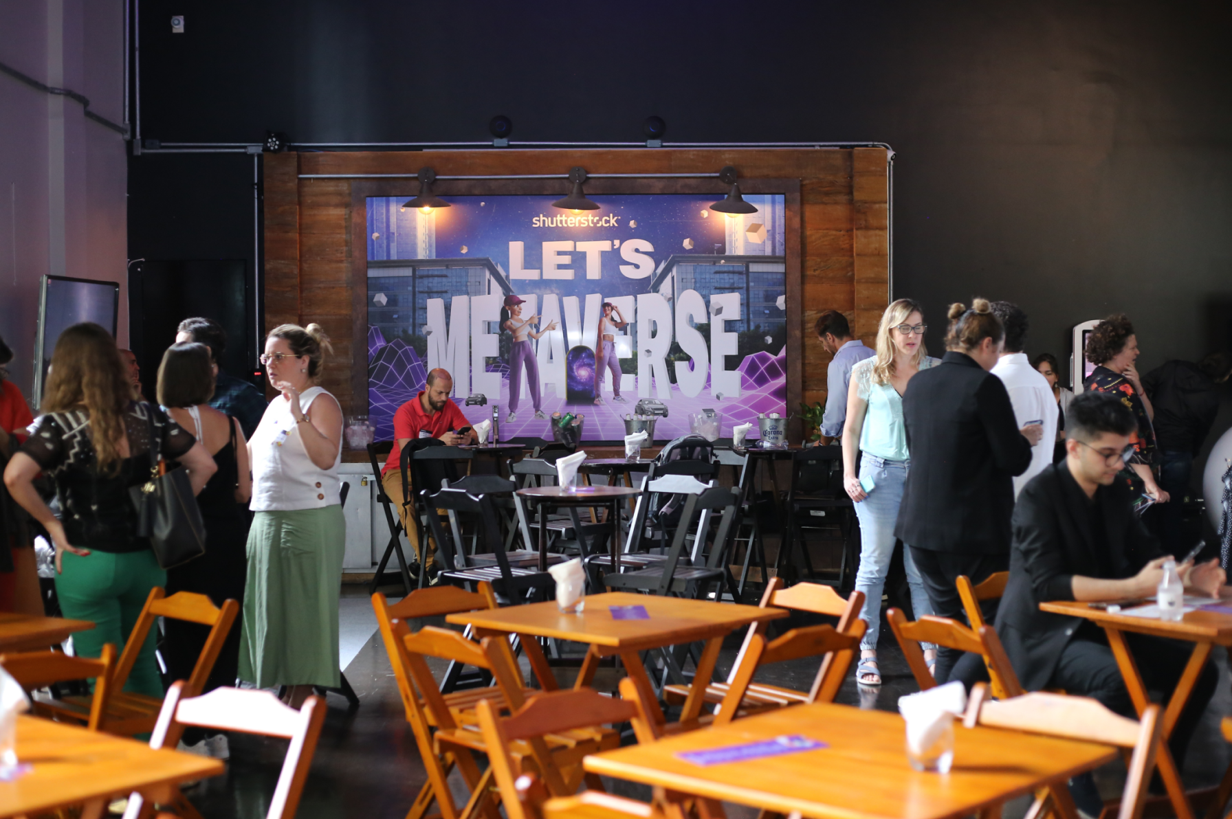 Indoor event with people standing and sitting, a stage with a digital backdrop reading 'Let's Metaverse', some tables and chairs, and a man sitting in the foreground looking at his phone.