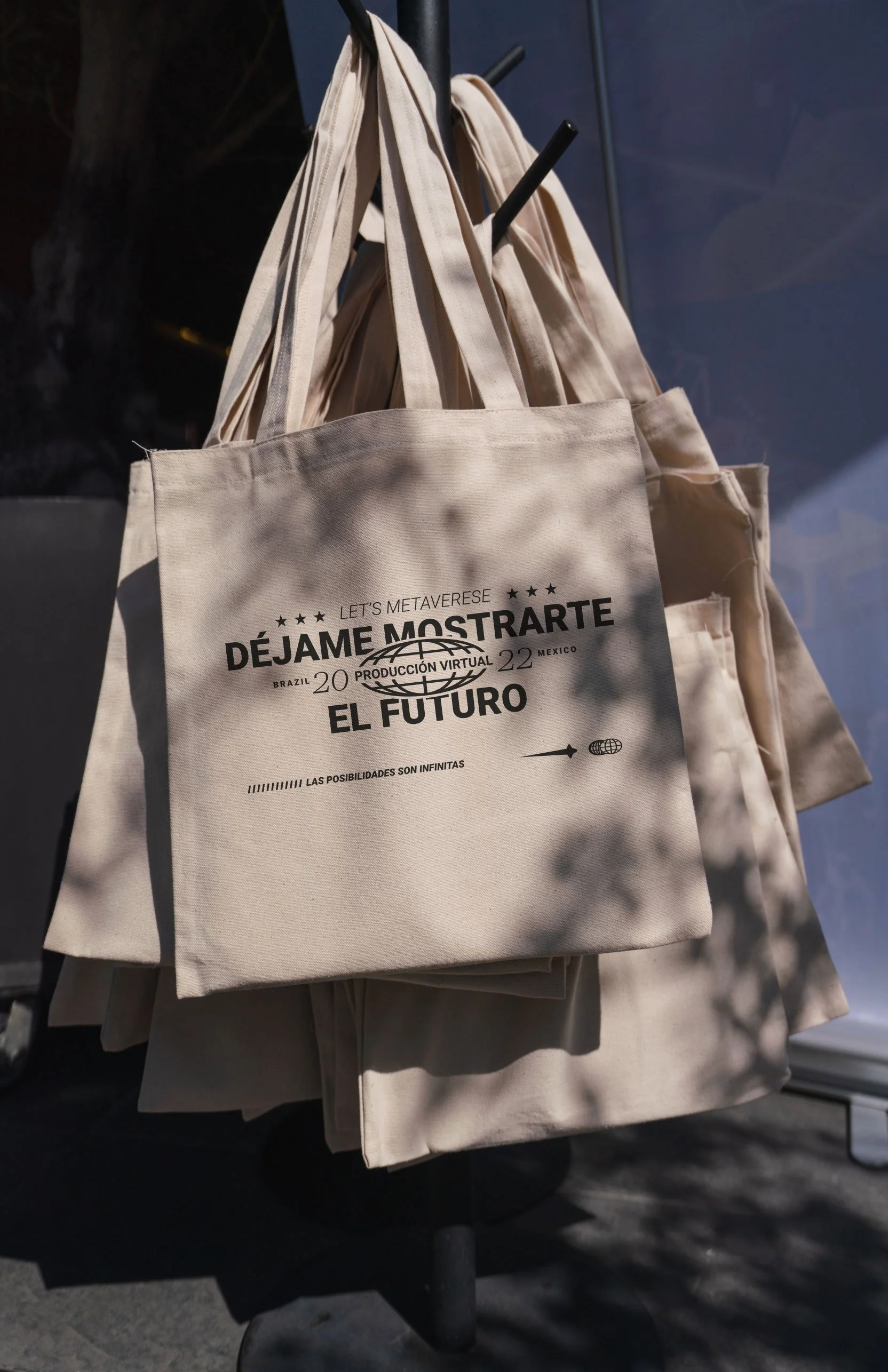 A beige tote bag hanging on a display stand, featuring black text and graphics promoting a virtual event about the metaverse, with the phrases "Let's Metaverse," "Déjame Mostrarte el Futuro," and other details in Spanish and English.