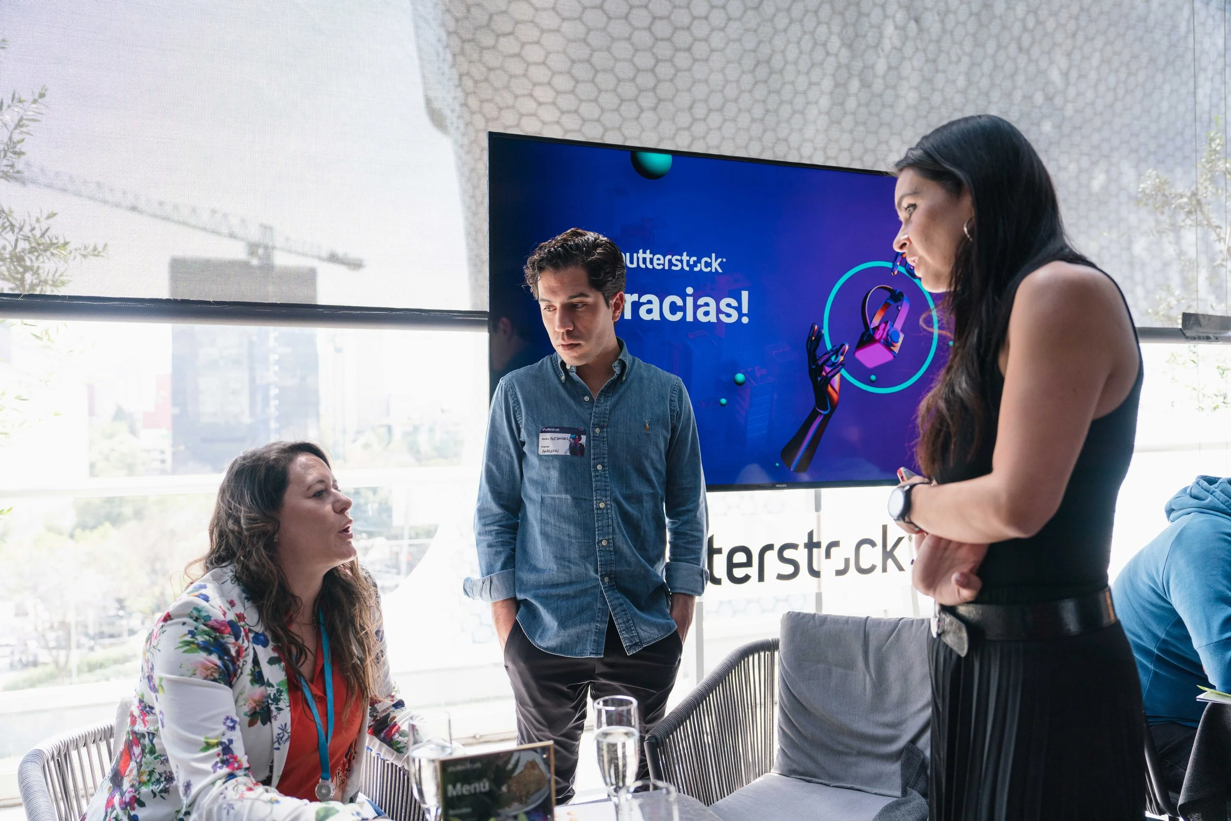 Three people in a business meeting or conference room having a discussion, with a large screen displaying thank you message and logos in the background.