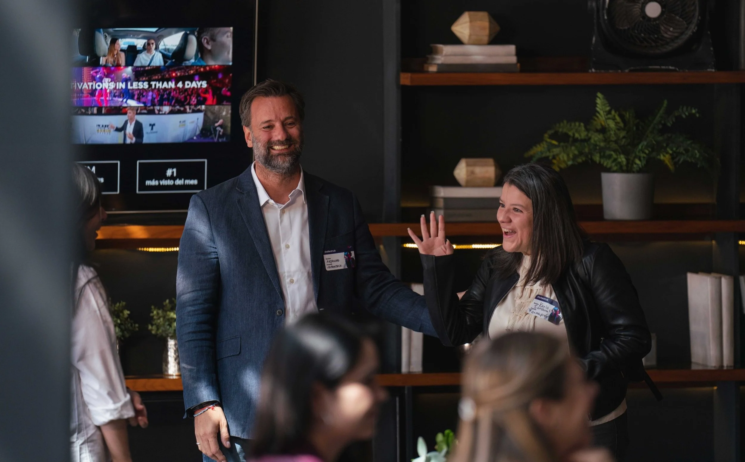 Two adults smiling and laughing at a social event, with women in the foreground, women on the left with glasses and blonde hair, woman on the right with dark hair and a leather jacket, man in a blazer and white shirt in the background, in a modern room with shelves, plants, and a television screen.