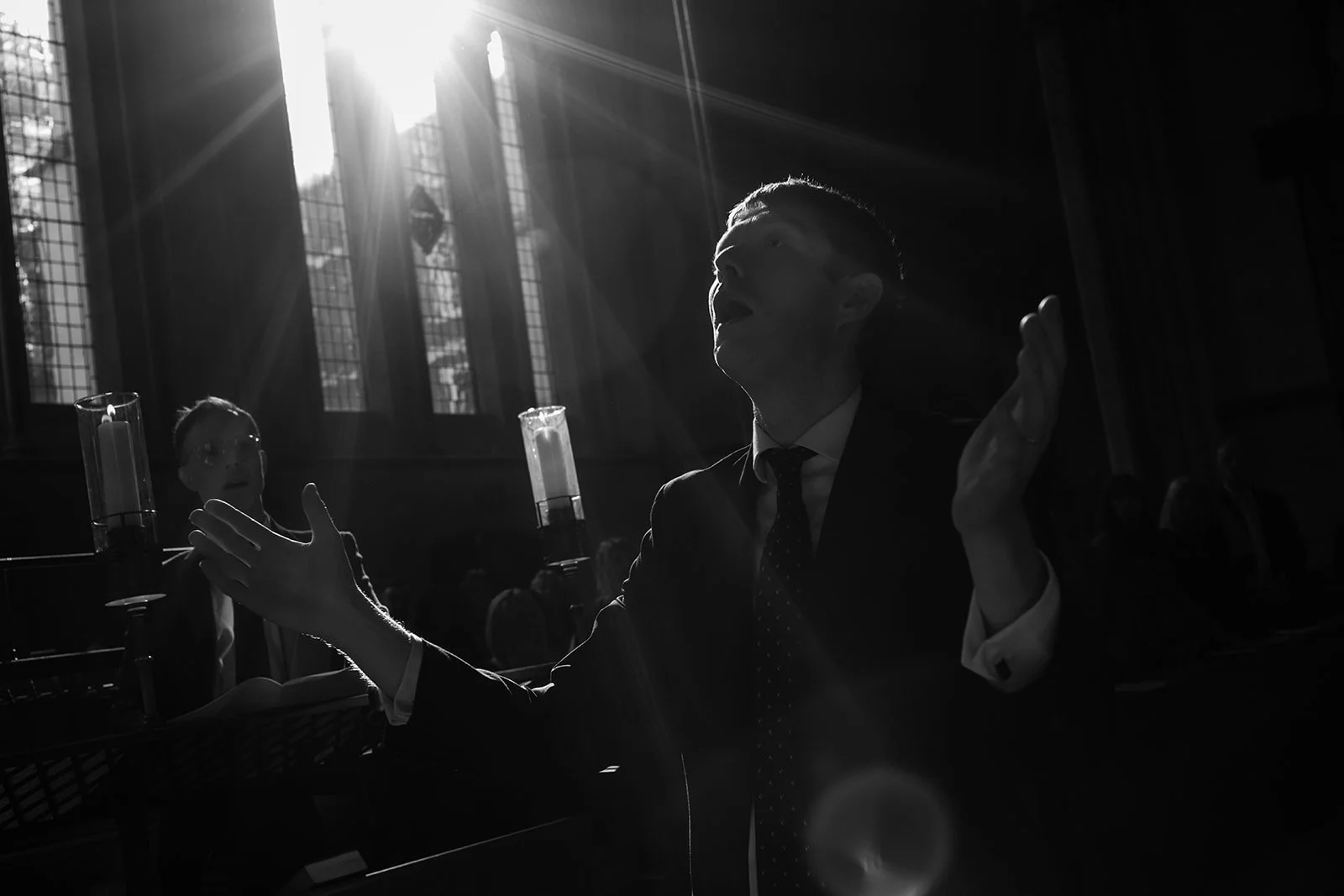 A man in a suit with a tie, raising his right hand, appears to be speaking or singing, in a dimly lit church with sunlight streaming through tall stained glass windows. A woman in the background is also visible, holding candles.