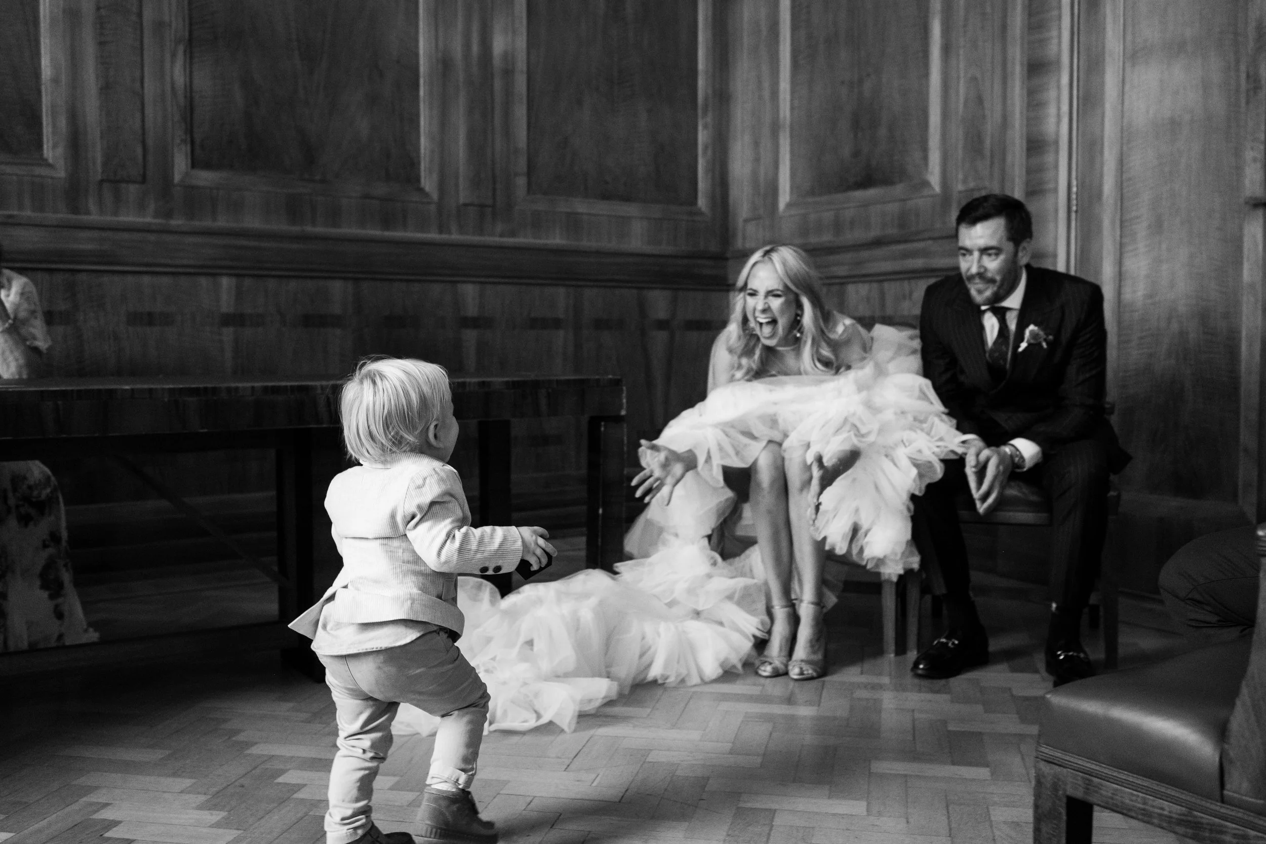 A young child in a suit happily interacting with a laughing bride in a wedding dress and a groom in a suit in a wood-paneled room.