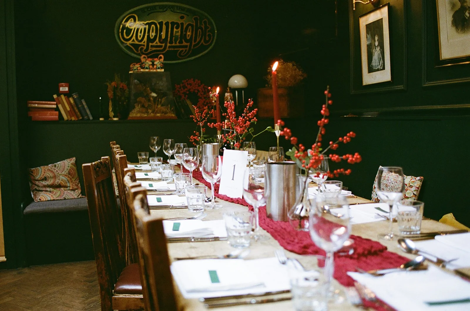 A long dining table decorated for a special occasion with a red table runner, floral centerpieces with red berries, tall red candles, and place settings including glasses, napkins, and utensils in a dark, cozy room with framed pictures on the wall.