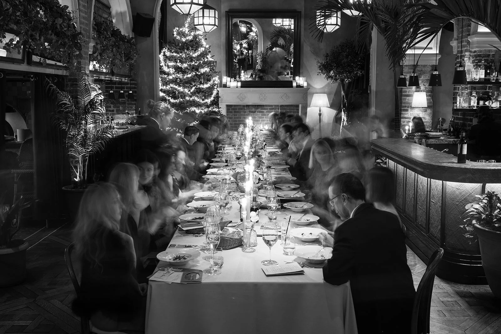 A group of people seated at a long dining table during a dinner event in a decorated restaurant with Christmas trees and candles.