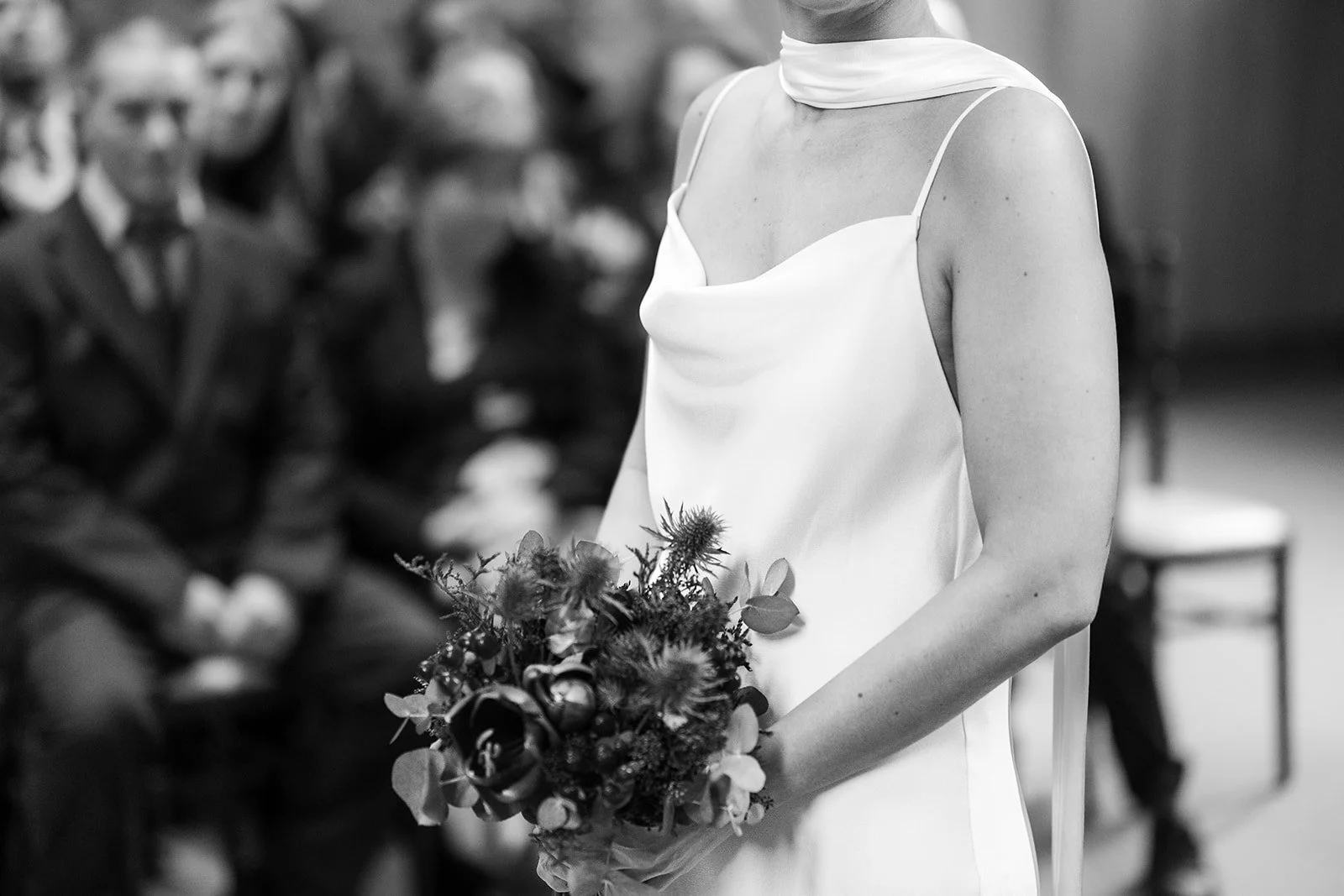 A woman wearing a white dress holding a bouquet of flowers at a wedding or formal event, with seated guests in the background.