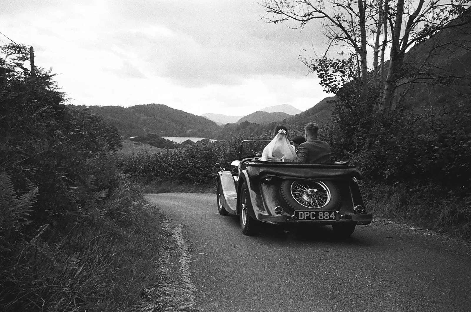 A black-and-white photo of a vintage convertible car driving on a rural road with a bride and groom sitting in the back seat, surrounded by hills and trees.