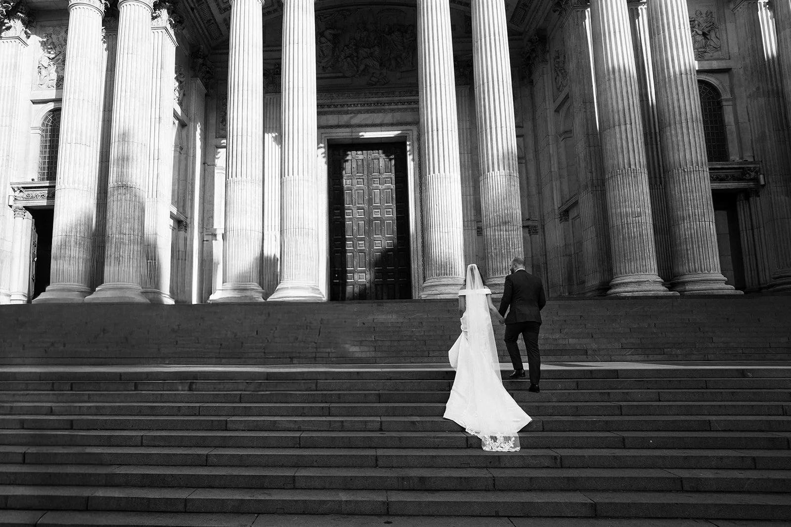 A black and white photograph of a bride and groom walking up the steps of a large, classical-style building with tall columns and ornate architectural details.