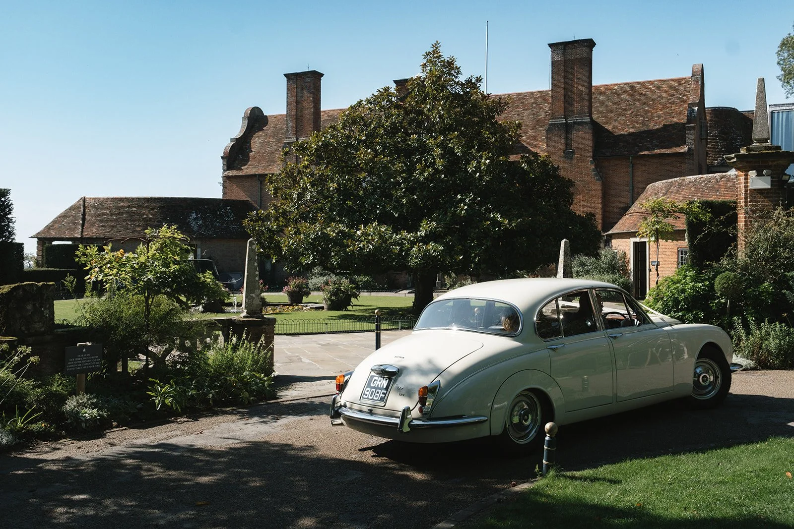 A vintage white car parked in front of a historic brick building surrounded by a garden and trees.