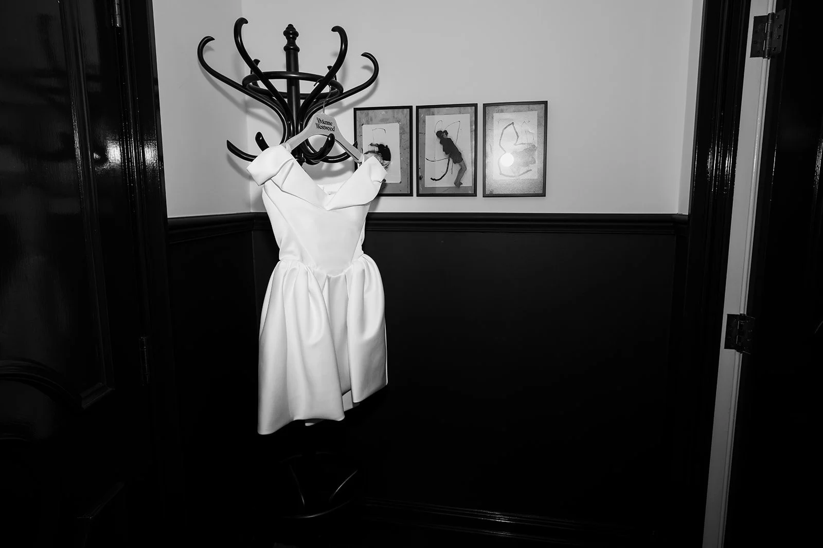 A white dress hanging on a black coat rack in a room with black and white framed artwork on the wall.