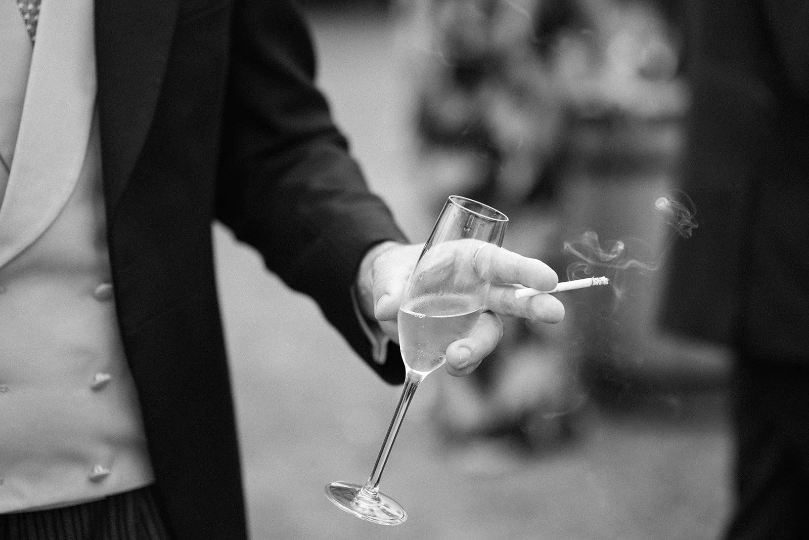 Person holding a champagne flute with one hand, with a lit cigarette in the same hand, in black and white.