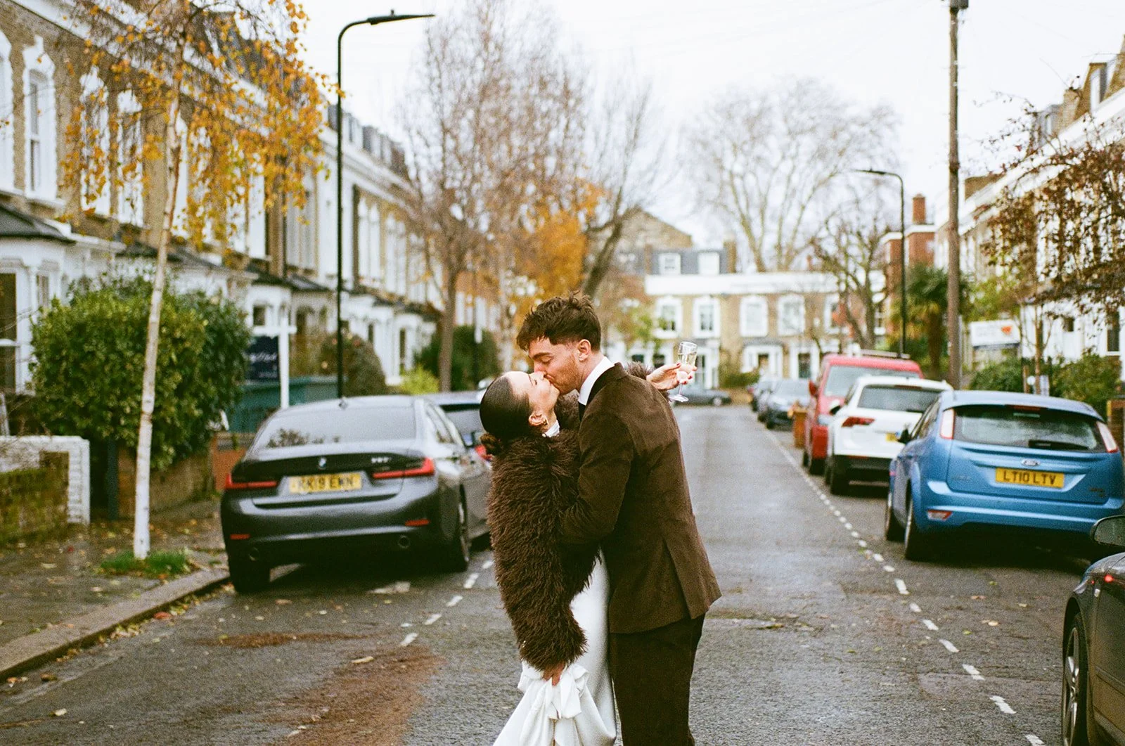 A couple is standing on a city street, sharing a kiss, with the woman in a white dress and fur coat and the man in a dark suit, holding a glass of champagne. The street is lined with parked cars and trees with autumn leaves.