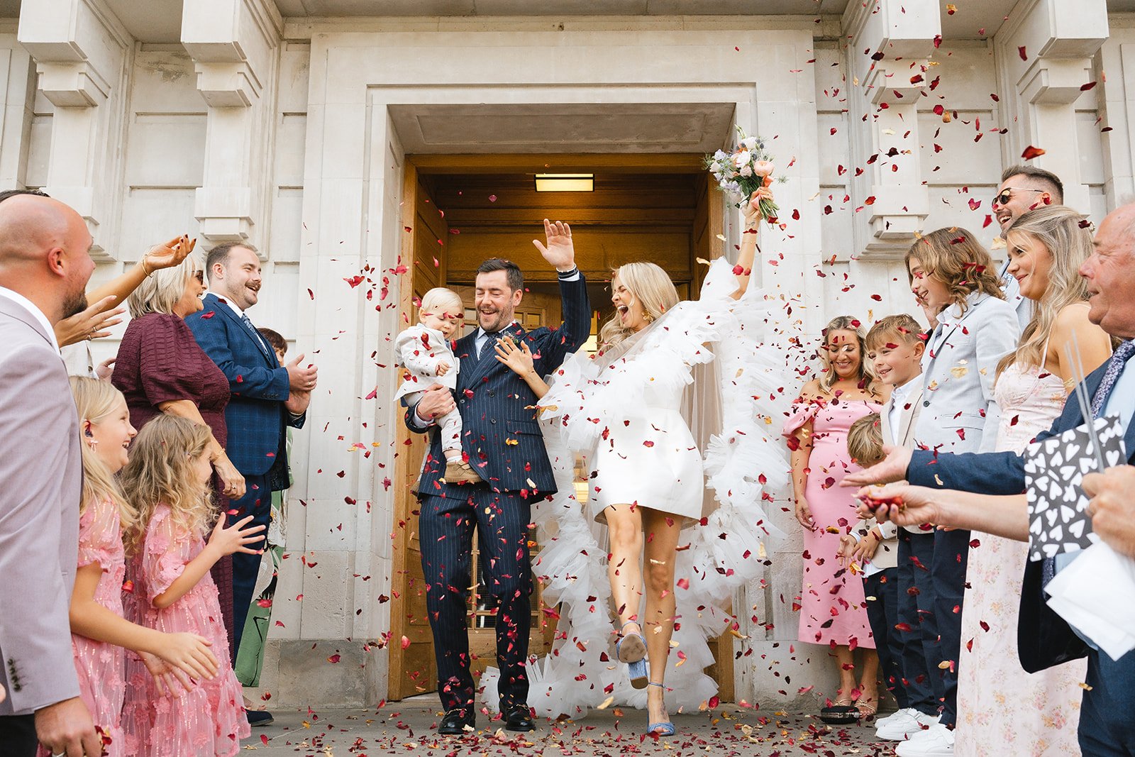 A newlywed couple celebrating their wedding exit with family and friends outside a church, surrounded by colorful confetti and flower petals.